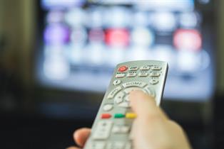 A remote control caught in shallow depth of field is being thrust at an out of focus TV screen