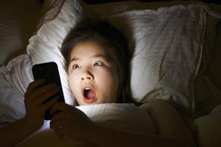 A teenager in bed appears shocked by something on their mobile phone, which we can't see