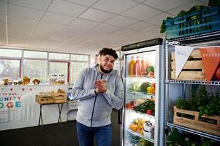 A volunteer from Feltham's Community Fridge smiles at the camera. The background is of the community fridge itself, with a fridges and shelves filled with vegetables. 