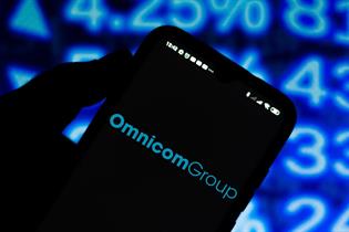 Omnicom's logo pictured on a mobile phone, in front of a stock market ticker display