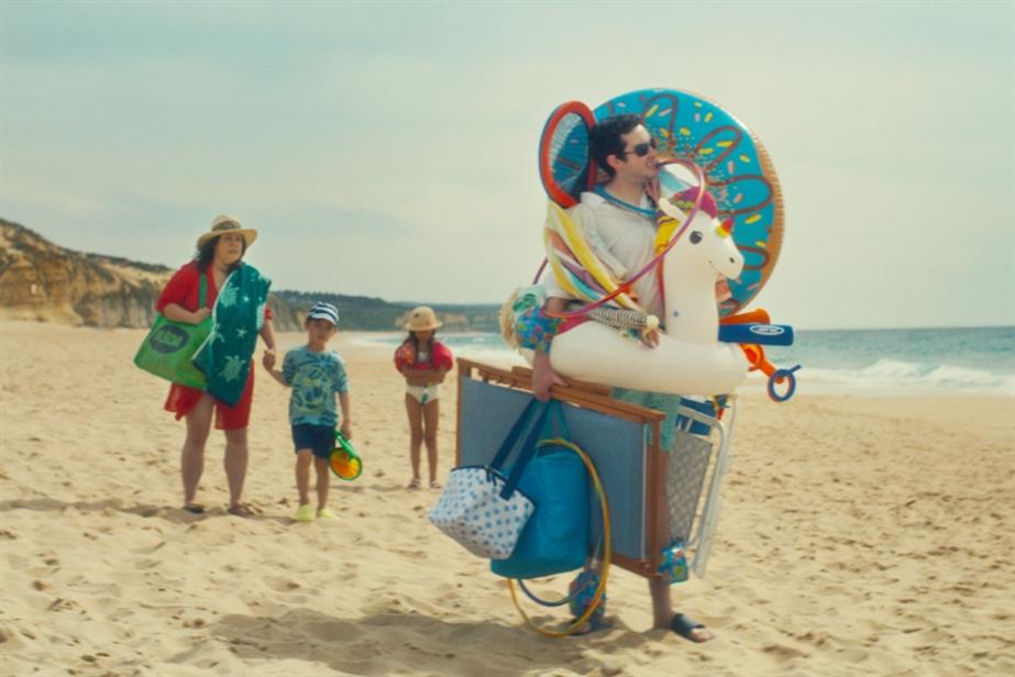 A family stand on the beach. The father is carrying a number of items as he searches for the perfect spot while his kids and wife trail behind. His wife is carrying an Asda bag.