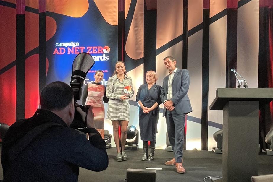 Three women accept an award onstage. A man, the presenter, stands slightly apart from them. 