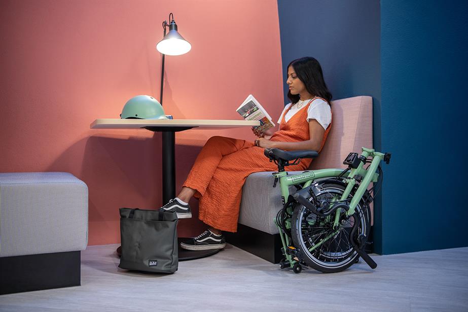 A girl reads a book on a chair next to a folded green Brompton bicycle