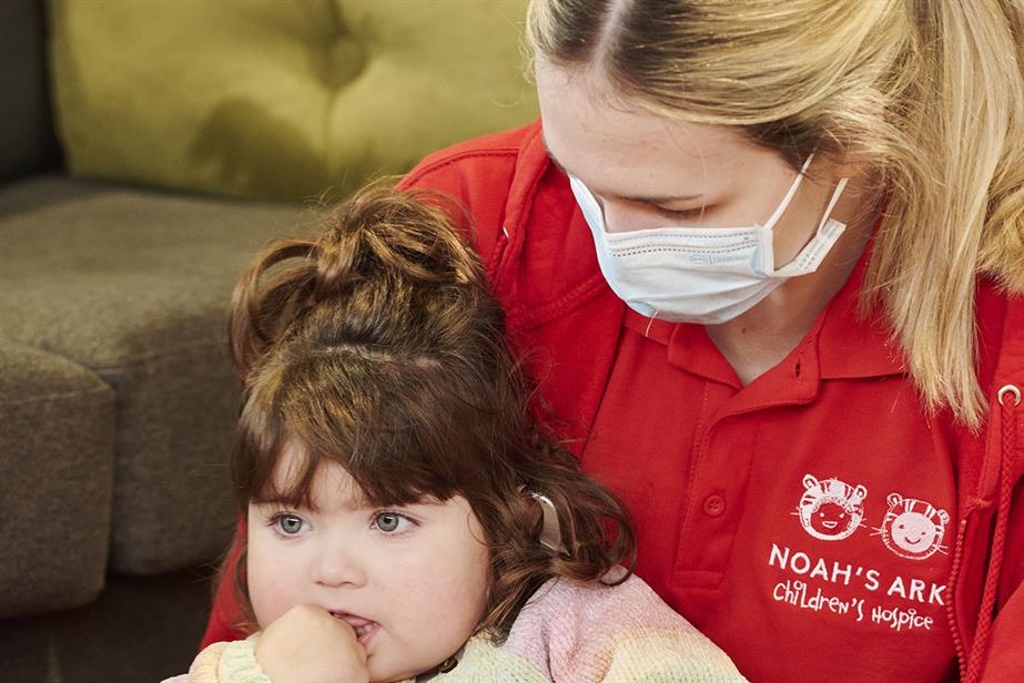 Picture of a female member of staff wearing a Noah's Ark Children's Hospice shirt and a mask hugging a little girl