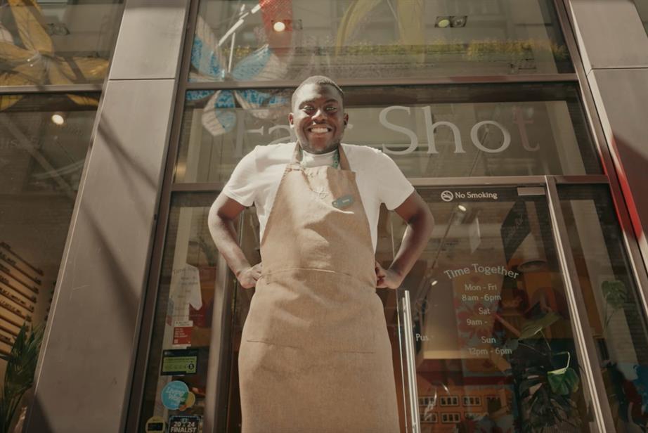 A black man stands outside the Fair Shot café, grinning down at the camera