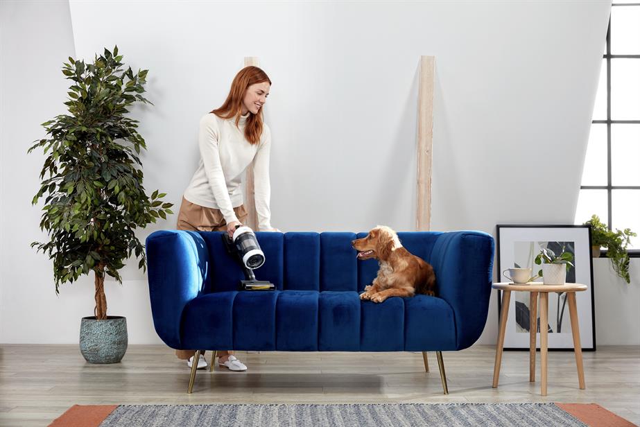 A woman hoovering her sofa while smiling at her dog