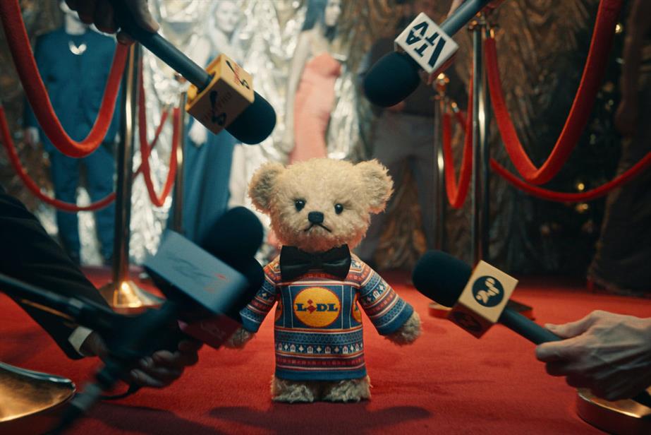 A teddy bear in a Lidl jumper and bow tie on a red carpet with microphones thrust at him