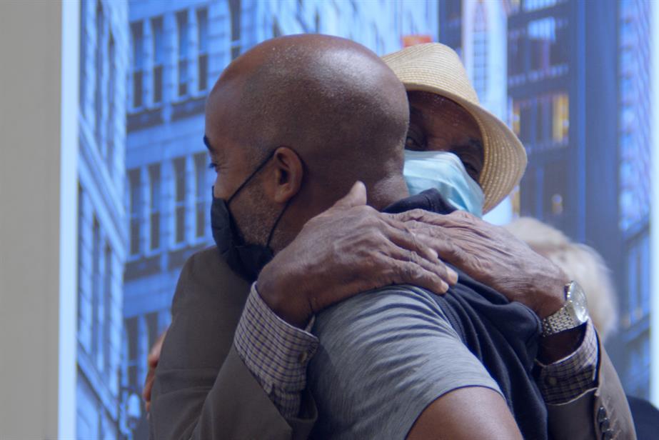 A black man and his older relative – both wearing Covid masks – hug each other as they reunite at Heathrow airport Arrivals