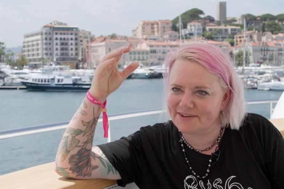 Woman with pink hair and tattoos smiles and gesticulates while talking, sat on a yacht in the harbour at Cannes