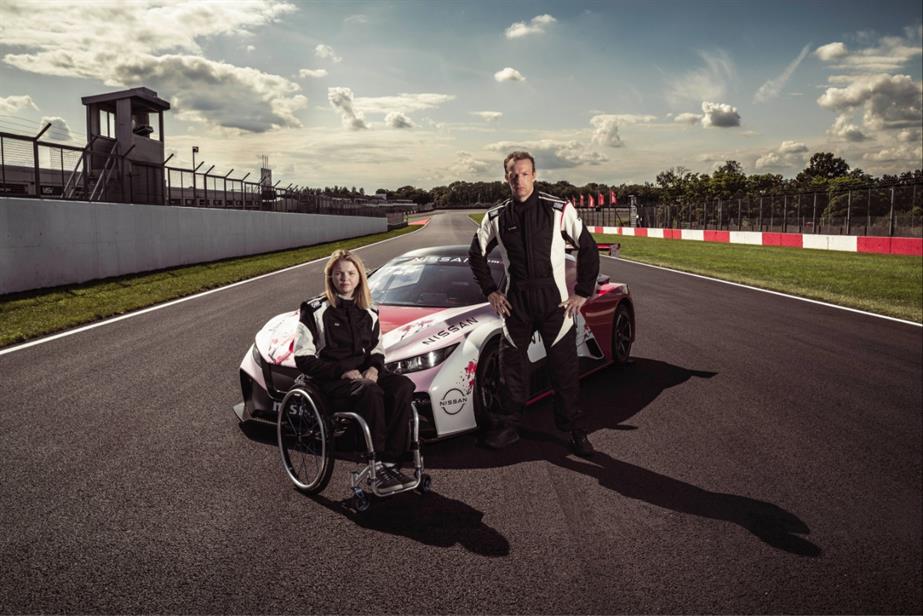A woman in a wheelchair and a man stand next to a car on a race track
