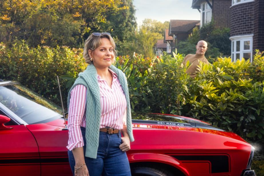 Woman smiling while stood next to a red convertible sports car as her neighbours peers over the hedge to look