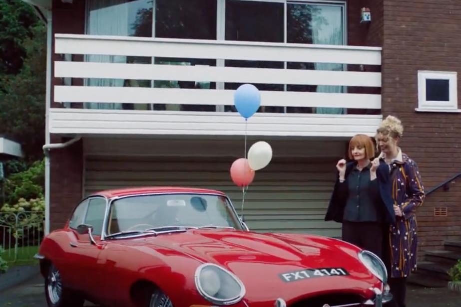 A mother guides a daughter to a brand new car. The daughter's hands are placed over her eyes and the car has balloons attached to it.