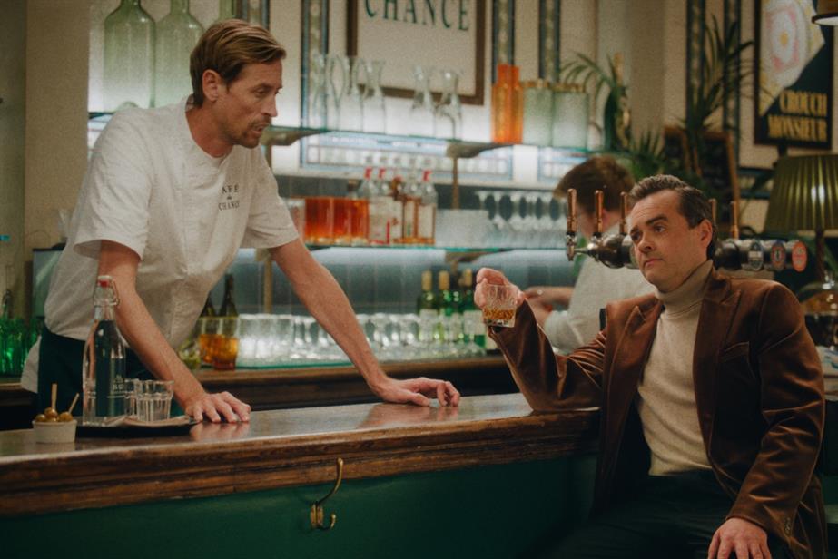 Peter Crouch stands behind a bar looking at a sat on a bar stool in front of him