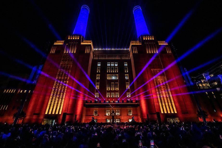 Battersea Power Station lit up at night with lasers coming out of it with a crowd in front of it