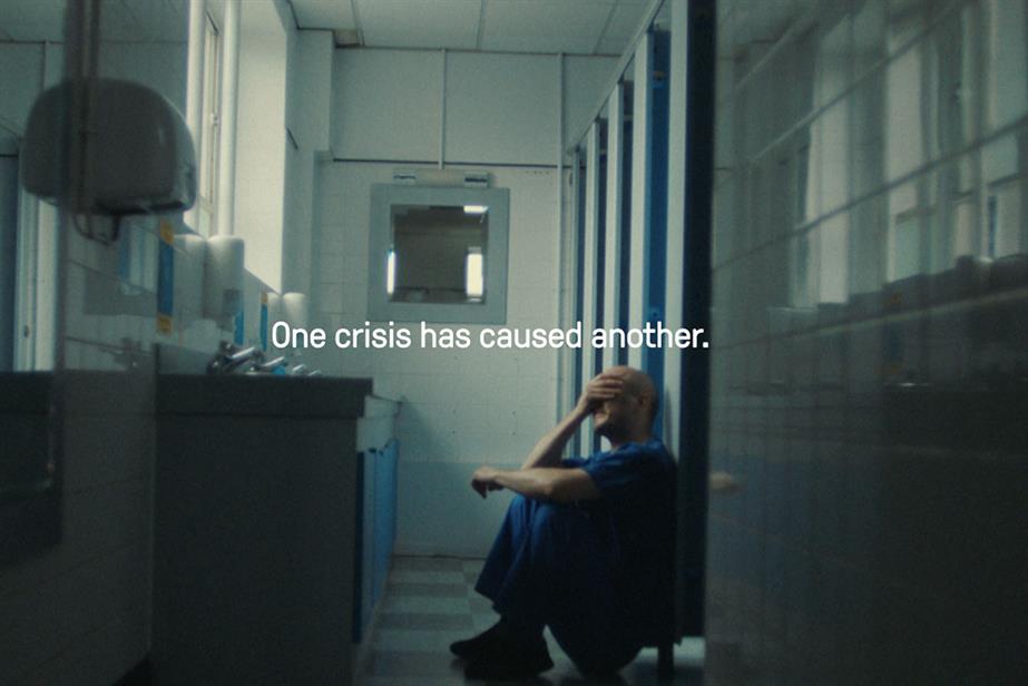 Image of a man in hospital scrubs sittting outside a row of toilet cubicles with his hand over his eyes and clearly in distress with the words 'one crisis has caused another' overlaid