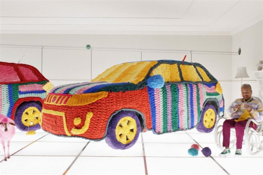 A Suzuki car covered in knitting with someone sitting in a chair knitting