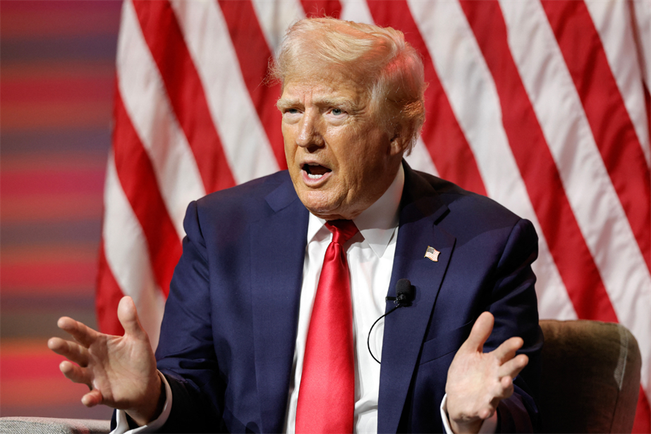 Image of Donald Trump's head and upper torso. He is seated in front of the US flat and is wearing a blue jacket, white shirt and red tie. His mouth is open, mid-speech, and his hands are lifted in front of his chest with palms open. 