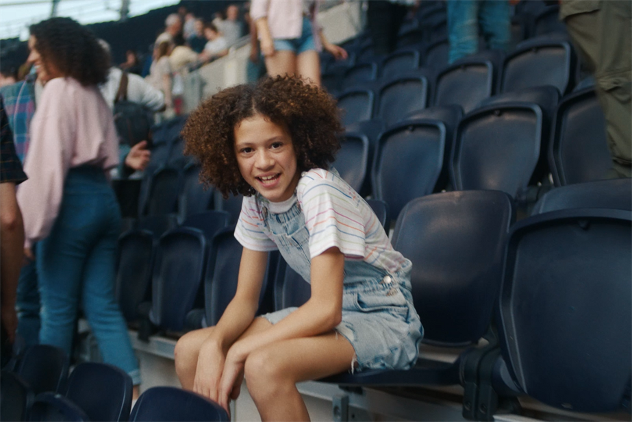 The girl protagonist sits in the stands after a Premier League game talking to the camera 