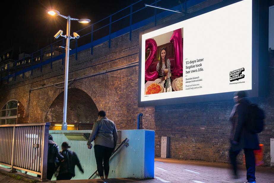 A billboard lit up at night next to railway arches showing an image of a smiling woman in a restaurant with the text: "11 days later Sophie took her own life" and the CALM logo