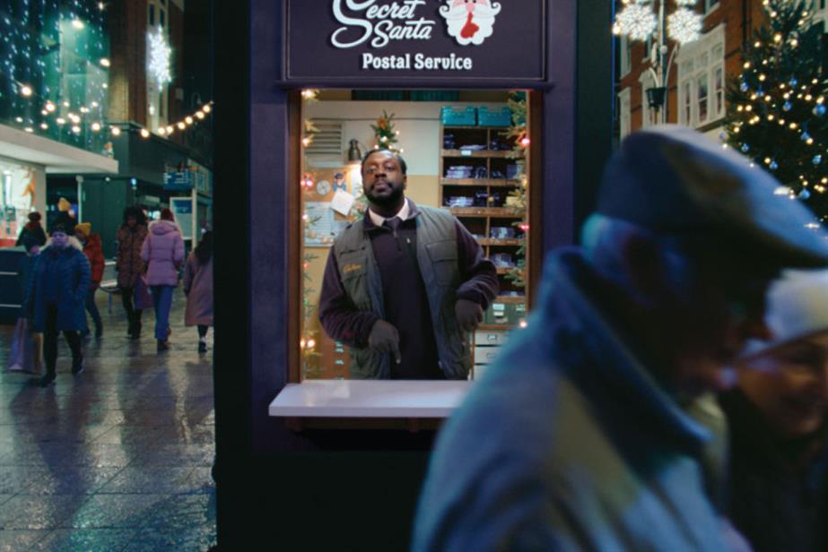 A ad for Cadbury with a man in a booth in the middle of a busy Christmas shopping street with a sign saying 'Secret Santa Postal Service' above him