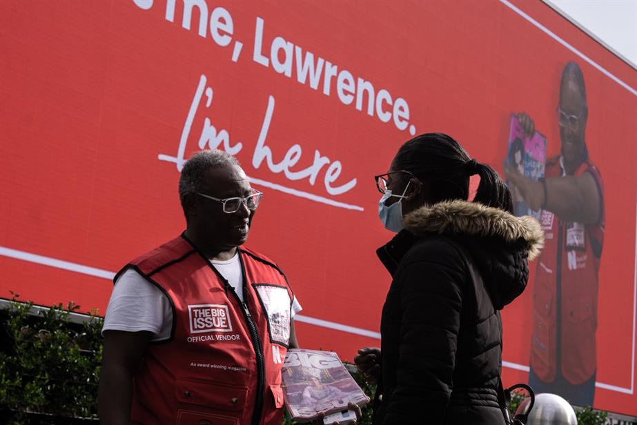 A Big Issue vendor called Lawrence chats with a woman buying a copy of the magazine, while a billboard in the background features a picture of him, saying 'I'm here' 