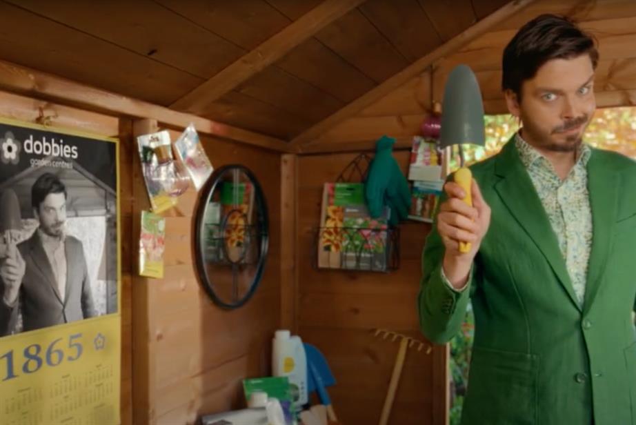 A man from Dobbies, wearing a green suit, stands in a shed, looking at the camera, holding a trowel aloft