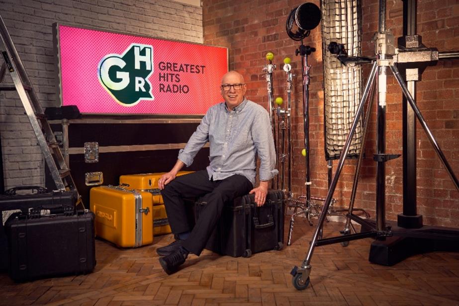 Ken Bruce sits on a chair in front of a TV with the logo for "Greatest Hits Radio" on.
