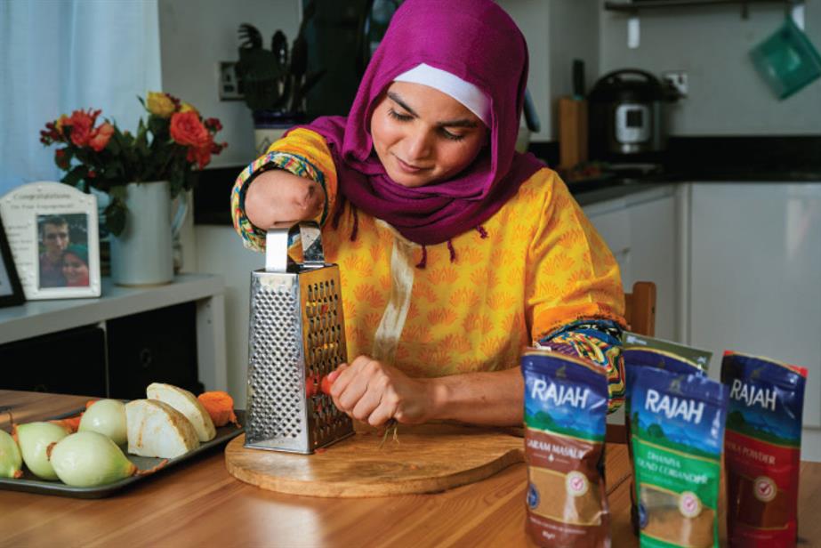 A woman in a headscarf grating carrot in a kitchen with Rajah spice packs on the table