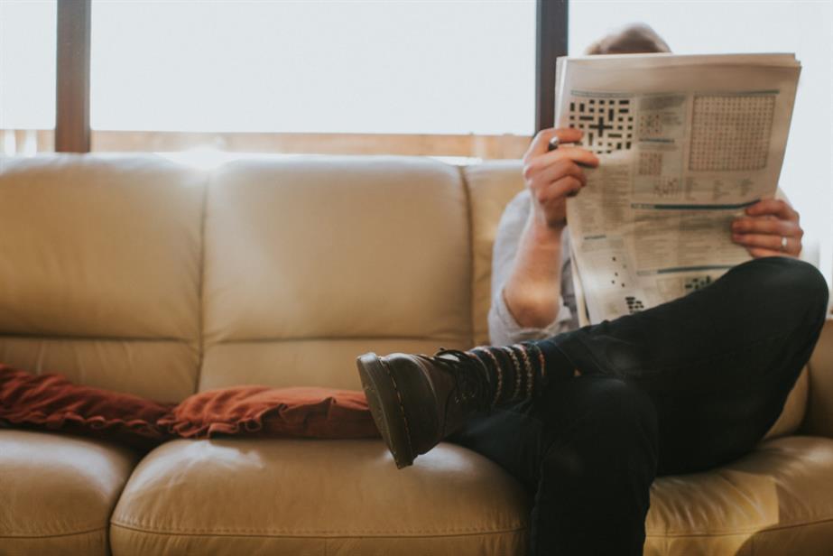 A man reading a newspaper at the end of a sofa