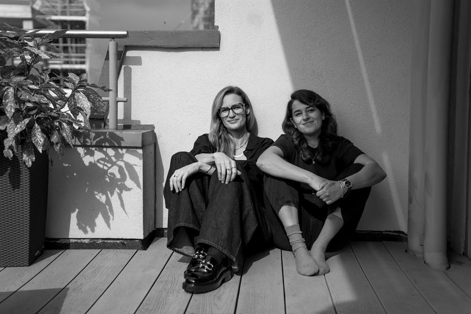 A black and white photo of Paula Bloodworth (left) and Sanam Petri, sitting on a wooden floor by a pot plant