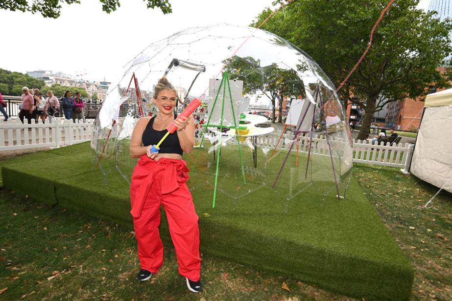 Liberty Poole attends Skittles' “The Goo Room”, dressed in red overalls and smiling at the camera. A clear dome with canvases is sat in the background. 