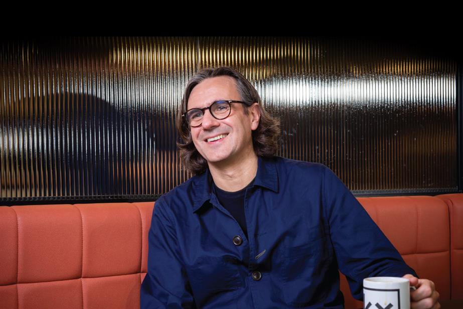 Head and shoulders shot of Xavier Rees sitting on a red banquette with a glass screen behind his head. He's wearing a blue shirt and specs and smiling