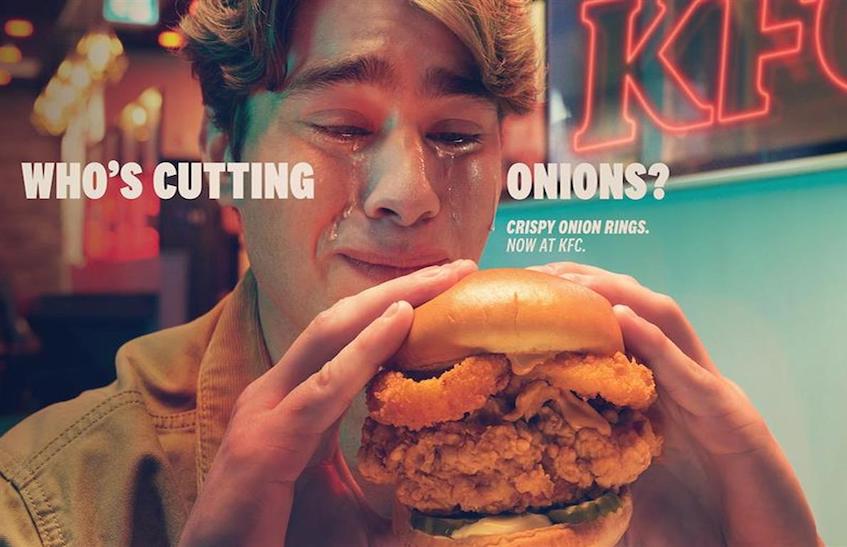 A KFC customer eating the new Crispy Onion Ring Sandwich while crying, the text reads "who's cutting onions?"