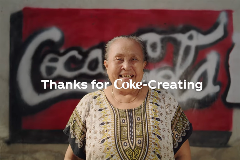 A woman smiles in front of Coca-Cola logo art with the slogan "Thanks for Coke-creating"