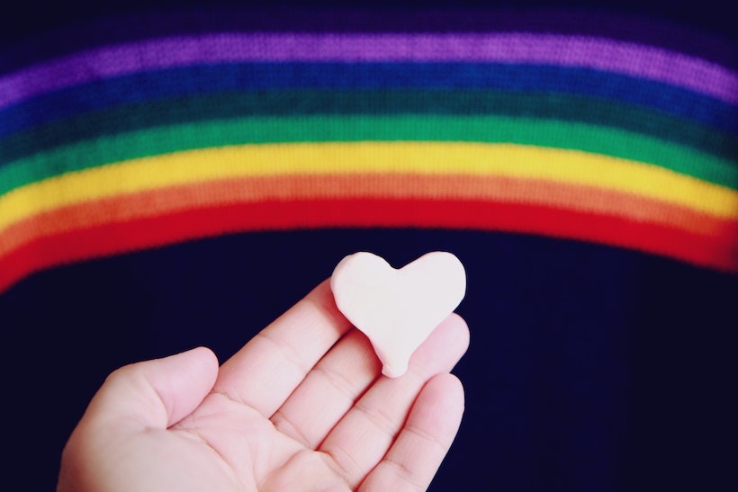 Hand holding heart in front of LGBTQ+ rainbow