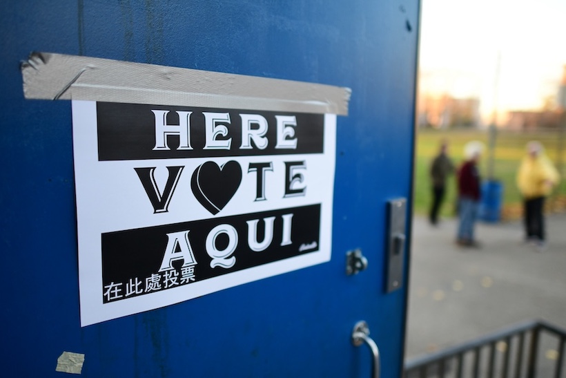 People stand outside a voting location that has a door with a sign reading "here, vote, aqui"