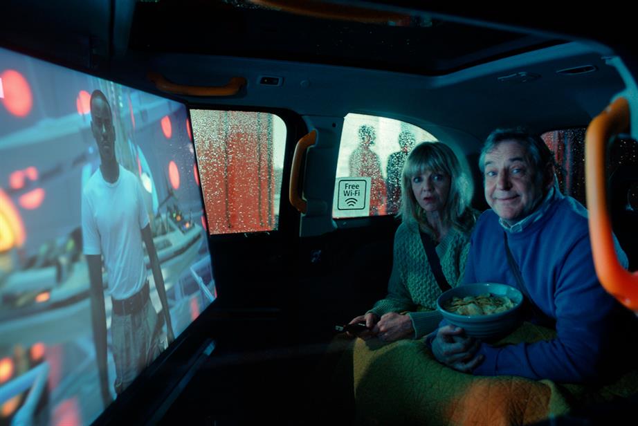 a middle aged couple sit in the back of a London cab, watching TV and eating snacks