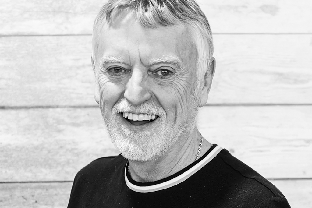 Black and white professional headshot of Clive Hyland, an older man with white hair and beard, wearing glasses and a black crew neck sweater with white trim, smiling warmly at the camera against a white wooden panel background