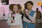 Two smiling children with Barbie-branded microphones smile at the camera with a film set behind them