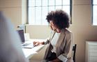 A confident businesswoman in a light-filled office, engaged with her laptop. She appears focused and professional, conveying a sense of productivity and modern work environment.