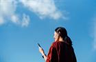 LOW ANGLE PORTRAIT OF YOUNG ASIAN WOMAN USING SMARTPHONE AGAINST BEAUTIFUL BLUE SKY WITH CLOUDSCAPES, ENJOYING SUNLIGHT OUTDOORS. LIFESTYLE AND TECHNOLOGY