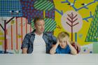 A small boy with his hands to his temples, sitting next to his dad at a table in front of a bright wall mural