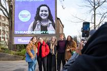 A group of people stand smiling in front of a billboard. On the billboard a smiling young woman and a circular logo with the words "Sign up now", and at the bottom of the poster, 'TheADcademy.org'.