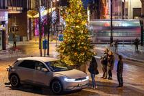 An electric car stopped next to a decorated Christmas tree in Glasgow city centre. Passersby are standing looking up at the tree.