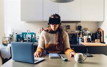 A woman wearing a VR helmet sits at her kitchen table with an open laptop on the side