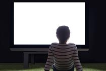 A girls sits in a dark living room in front of a large TV bearing a blank white screen