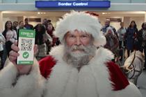 Santa is seen standing in the foreground at an airport, holding his smartphone up so that viewers can see his Covid-19 Passport, while grinning travellers look on behind him