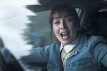 A woman faces the camera as she sits in her car. Mouth open and arms wide, she is shouting in anger.