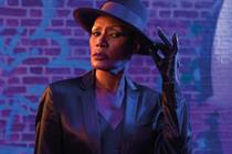 Grace Jones in a black hat, jacket and leather gloves standing in front of a brick wall