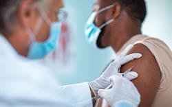 Blurred image of a black man wearing a mask being injected in he upper arm by a medical professional also wearing a mask